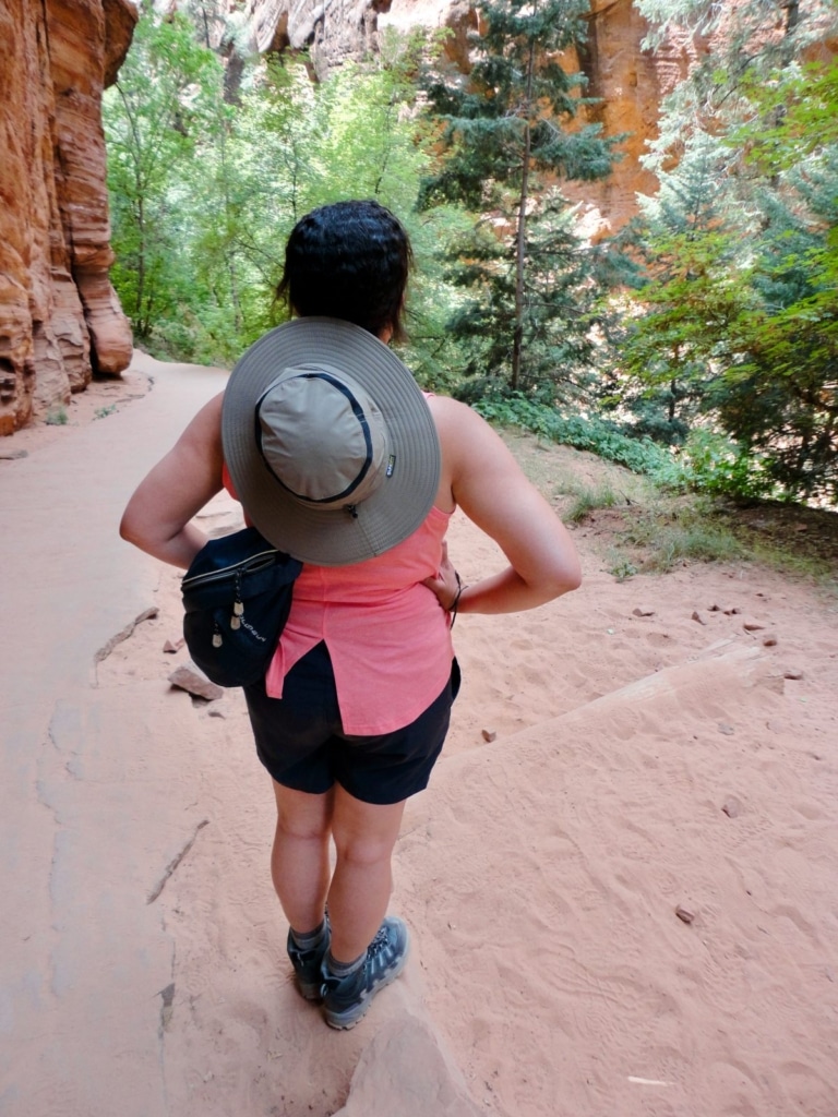 Woman with hiking clothes, hat, and boots standing on a trail with trees on the side