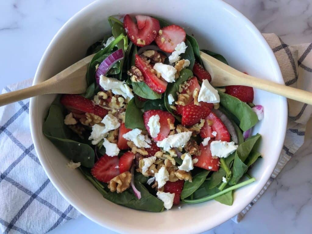 strawberries, red onions, spinach, walnuts, and goat cheese in a white bowl with 2 wooden spoons.