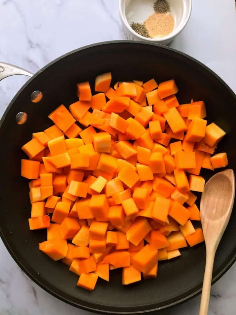 butternut squash cubes in a skillet with spices and wooden spoon.