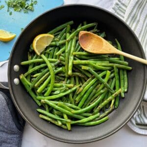 Green beans in a skillet with a lemon wedge and wooden spoon.